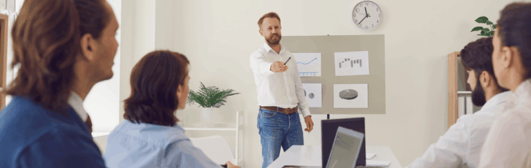 Bearded man in a white shirt presents data to four coworkers in a bright office, pointing at charts on a board